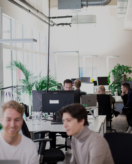 Modernes Großraumbüro mit mehreren Arbeitsplätzen, Mitarbeitenden an Computern, grüne Pflanzen und helle Einrichtung.