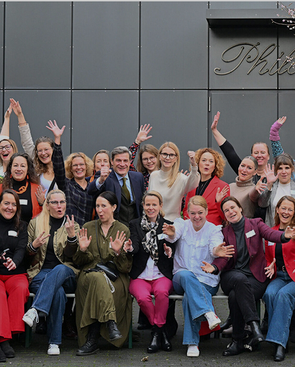 Gruppenfoto von etwa 25 Frauen und einem Mann in der Mitte vor grauer Wand, viele winken in die Kamera.