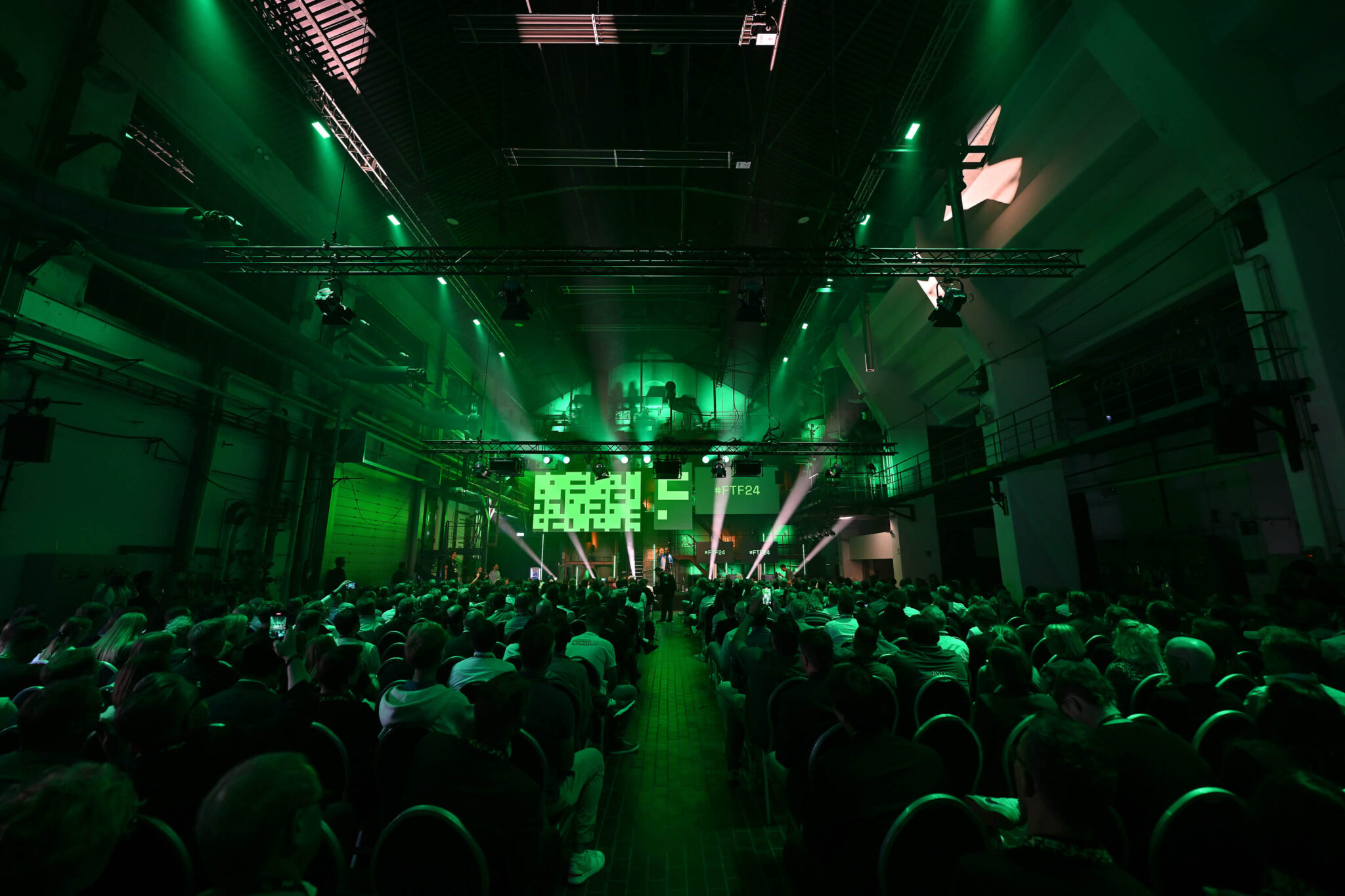 Volle Halle mit grüner Beleuchtung und hunderten Teilnehmern vor einer illuminierter Bühne.