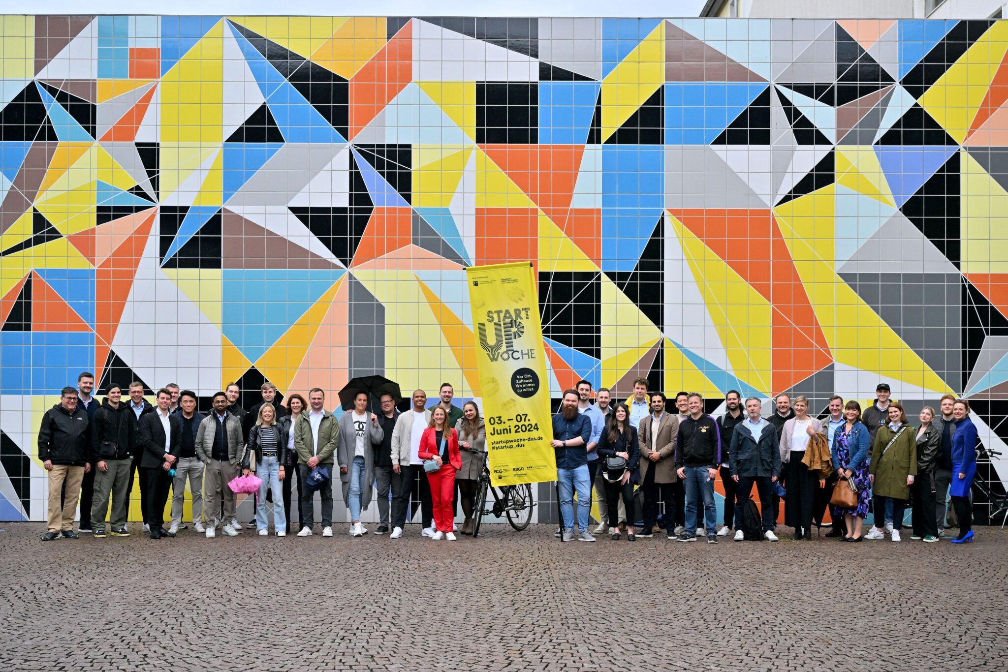 Startup Week Düsseldorf 2024 Gruppenfoto: Teilnehmende vor bunter Mosaikwand mit geometrischen Mustern in Gelb, Blau, Orange und Schwarz.