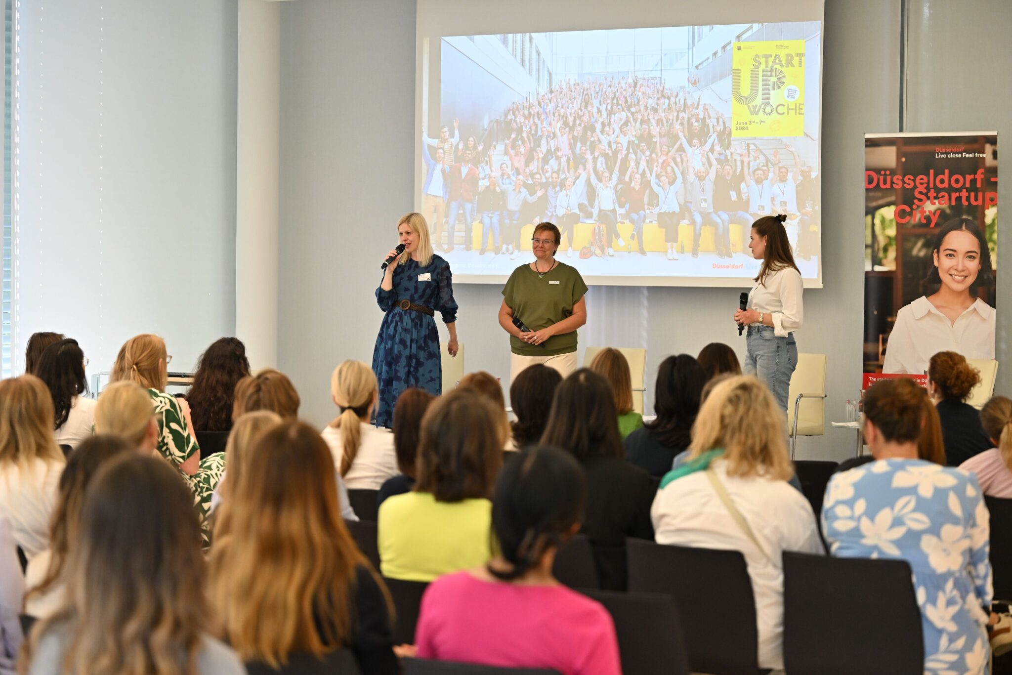 Startup Week Düsseldorf Panel-Diskussion: Drei Sprecherinnen präsentieren vor vollem Publikum, Gruppenfoto der Teilnehmenden auf Leinwand im Hintergrund.